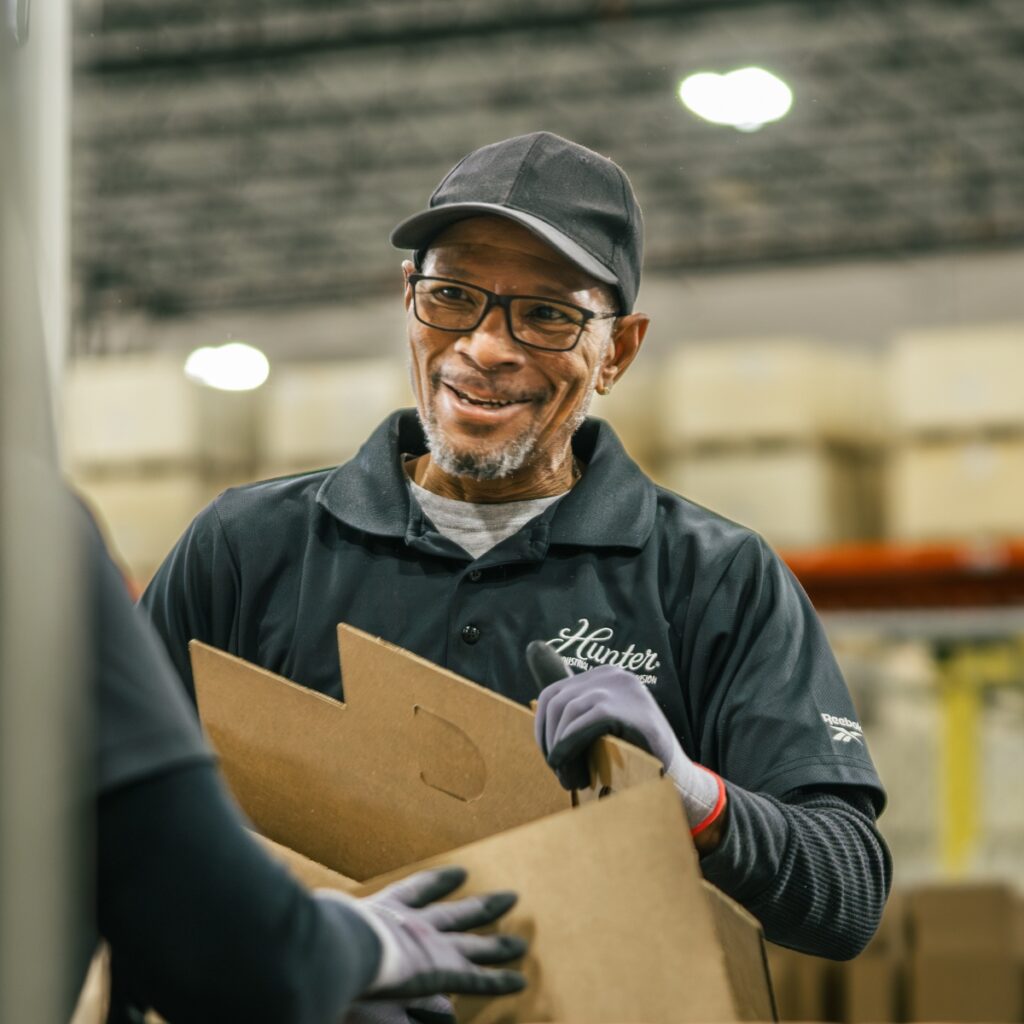 Opérateur logistique souriant portant un carton dans un entrepôt équipé de déstratificateurs